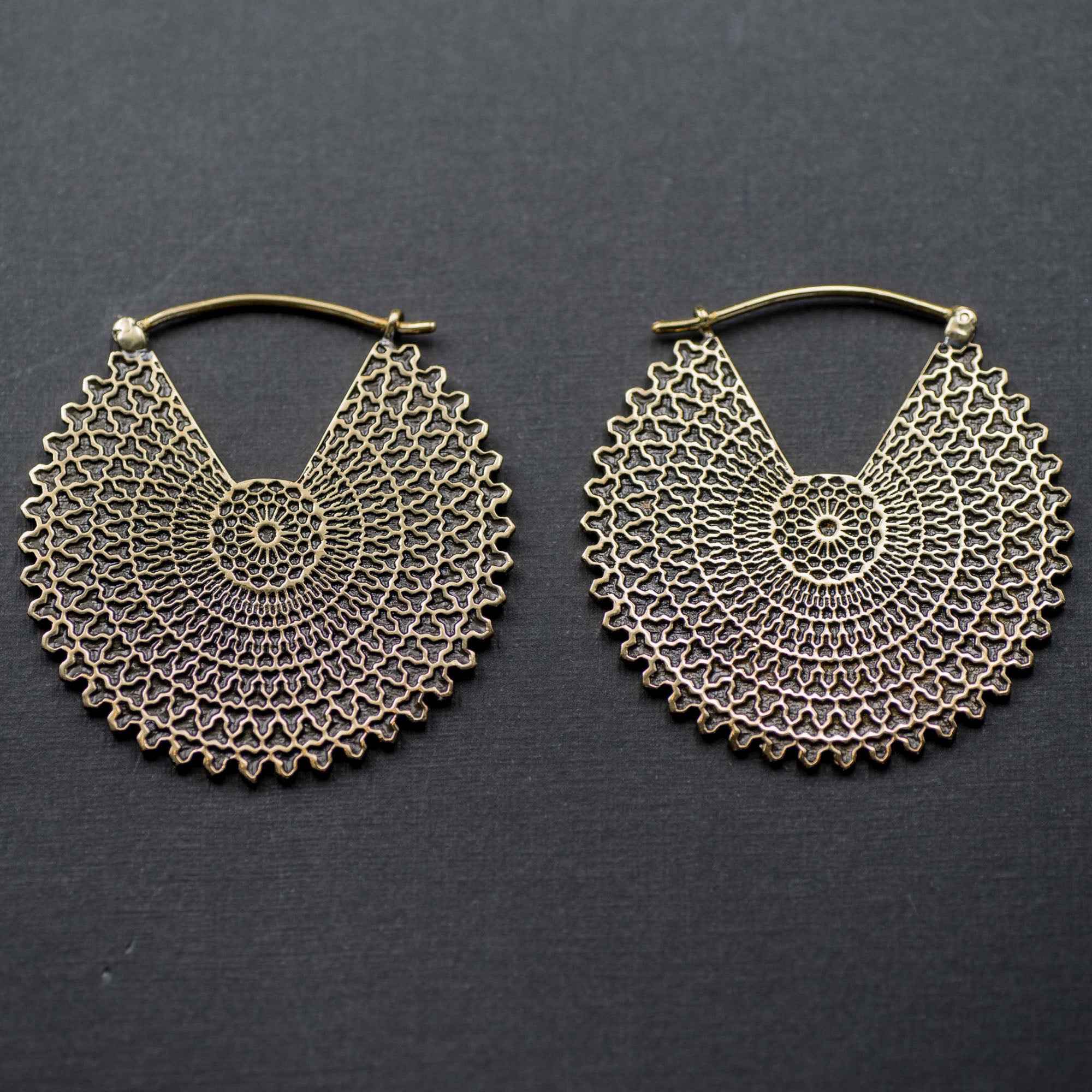 A pair of VORTEX Tribal Oversized Mandala Disc Hoop Earrings in gold, by Kaya Movement, showcasing an intricate latticework with concentric circles and a scalloped edge design, reminiscent of boho statement earrings, laying against a dark gray textured background.